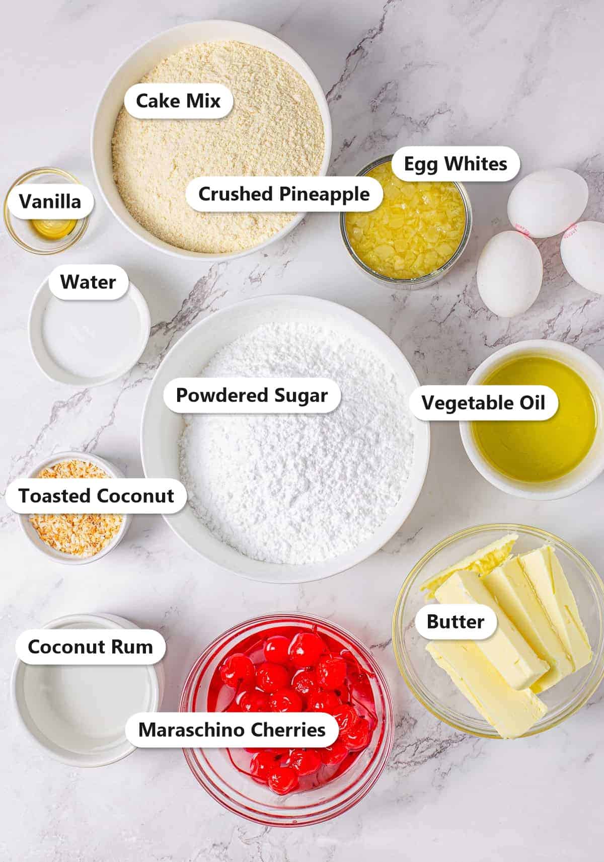 Pina colada cupcake ingredients labeled including cake mix, crushed pineapple, egg whites, vegetable oil, water, powdered sugar, butter, vanilla, coconut rum, toasted coconut, and maraschino cherries.