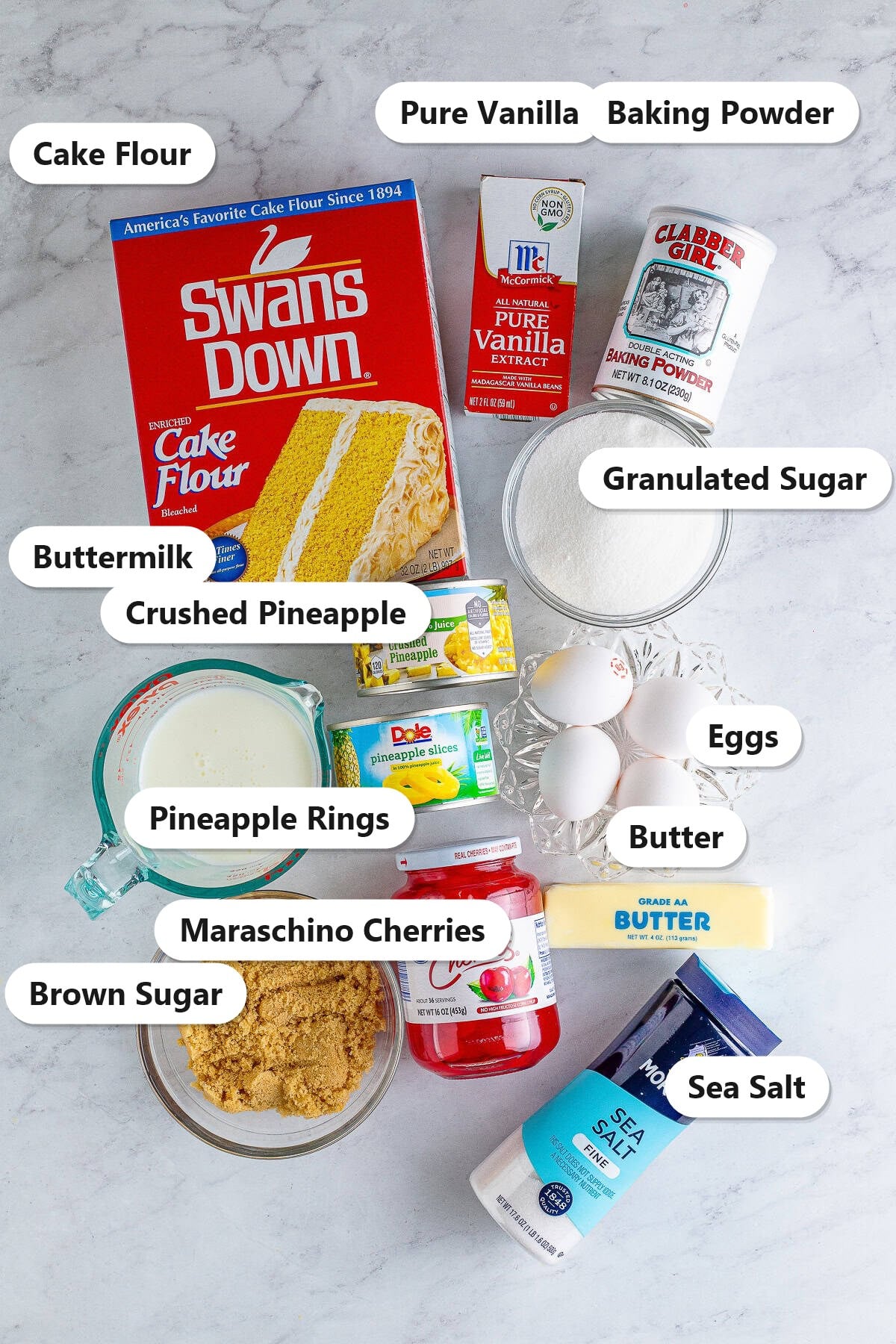 Pineapple Upside Down Cupcakes ingredients including cake flour, butter, sugar, eggs, buttermilk, pineapple, and cherries labeled on a counter.