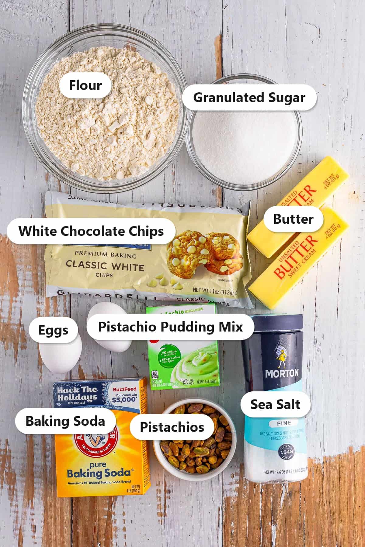 Labeled ingredients for pistachio pudding cookies including flour, pistachio pudding mix, butter, white chocolate chips, and shelled pistachios.