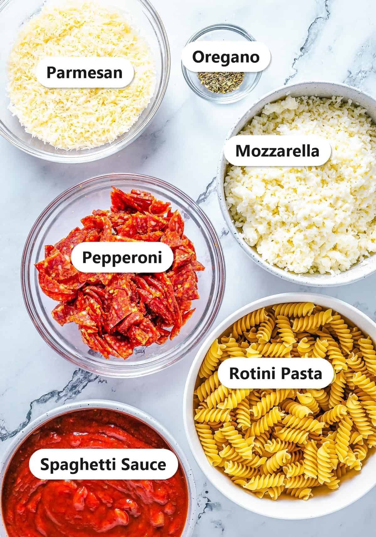 Pizza pasta casserole ingredients including rotini pasta, spaghetti sauce, mozzarella, pepperoni, parmesan, and oregano labeled on a marble surface.