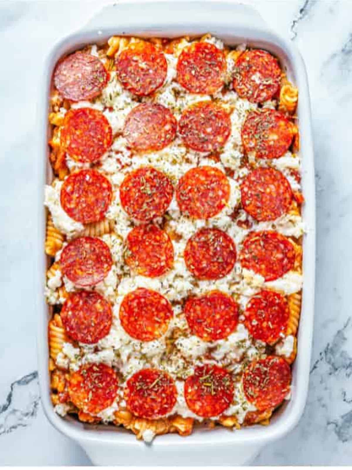 Pizza pasta casserole assembled in a baking dish and topped with pepperoni before baking.