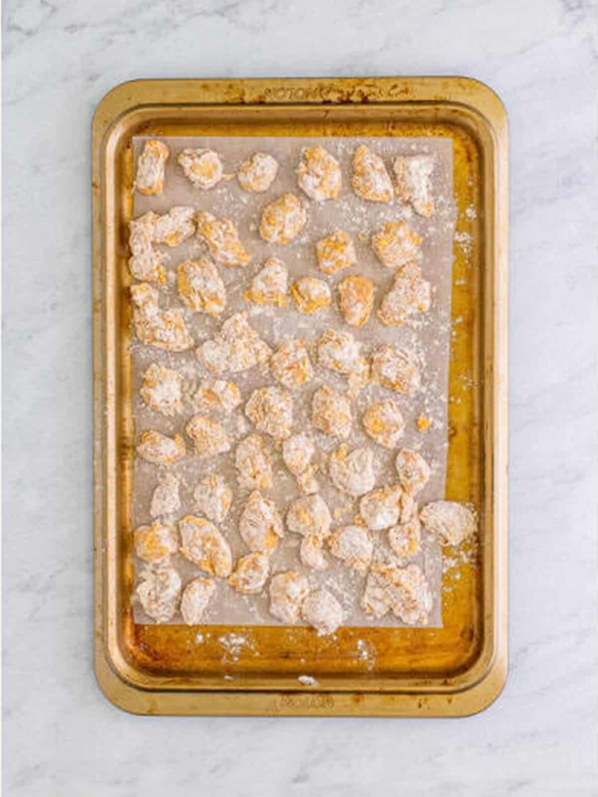 Breaded chicken pieces laid out on a sheet tray before frying for popcorn chicken.