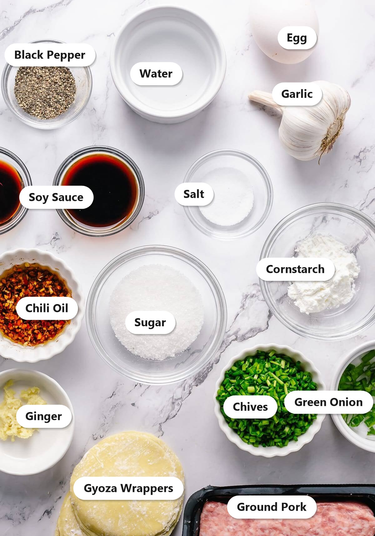 Ingredients to make pork dumplings labeled on a marble surface: ground pork, egg, chives, soy sauce, cornstarch, garlic, ginger, black pepper, salt, sugar, green onion, chili oil, water, and gyoza wrappers.