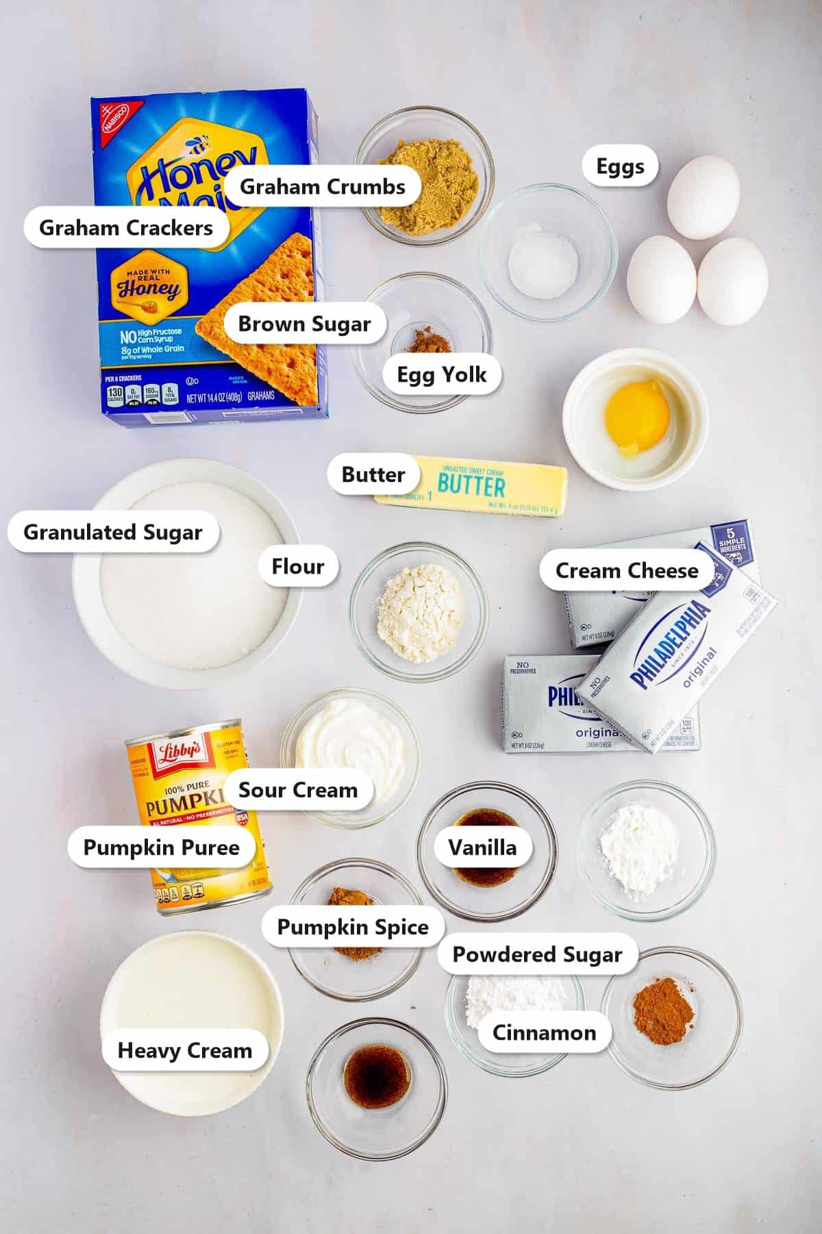 Pumpkin cheesecake ingredients labeled on a marble surface including graham crackers, cream cheese, pumpkin puree, eggs, sour cream, granulated sugar, butter, and pumpkin spice.