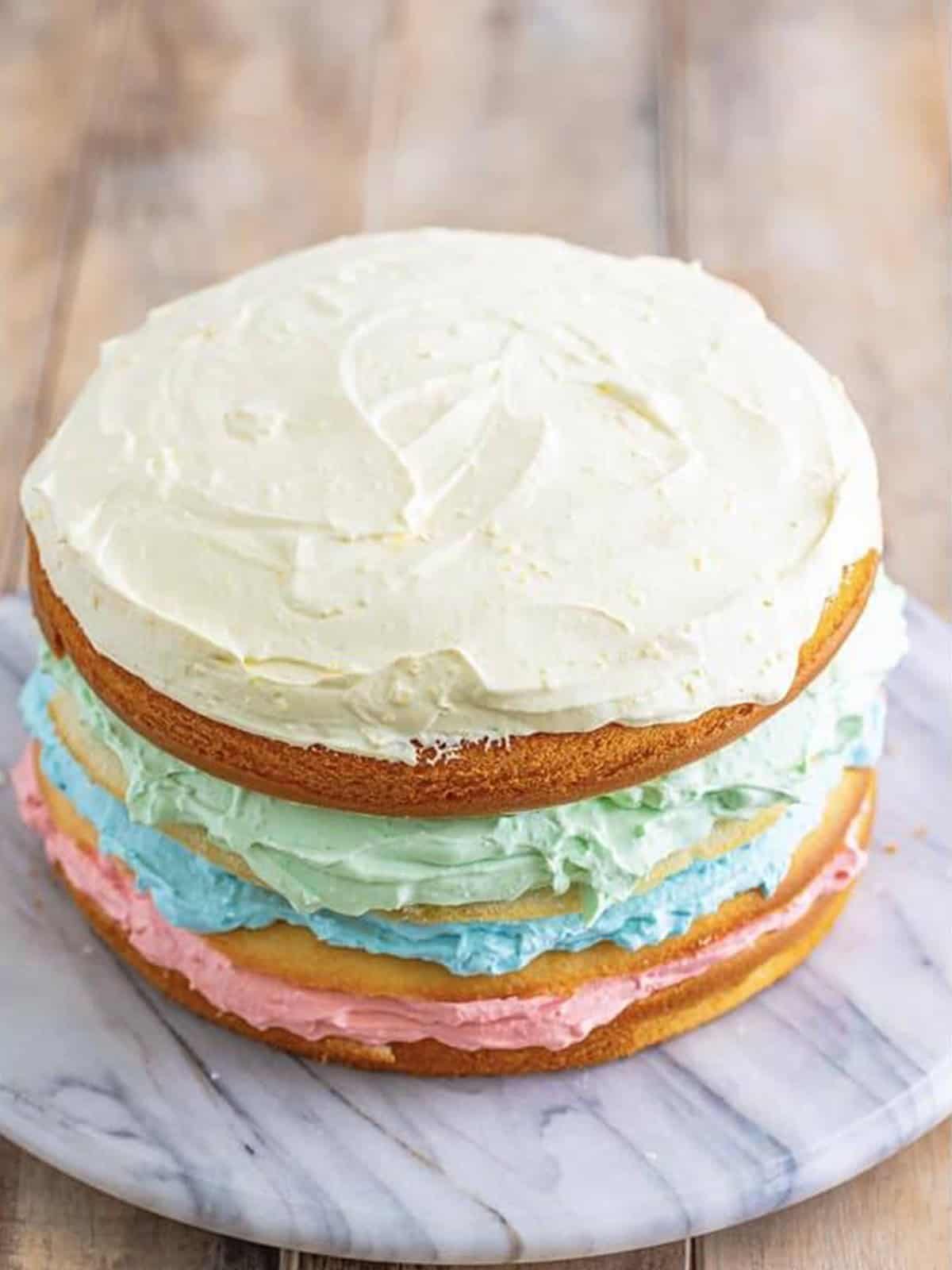 Stacked rainbow jello cake layers with pink, blue, green, and white frosting.