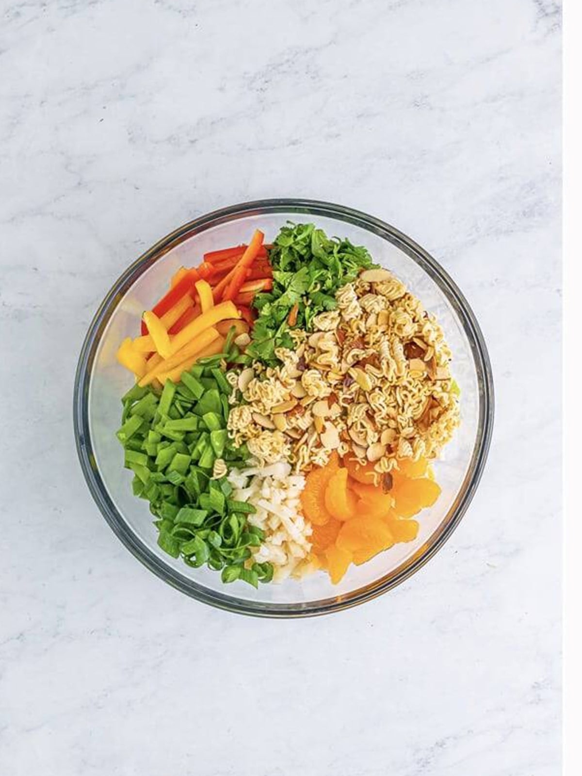 Coleslaw, bell peppers, snow peas, mandarin oranges, and toasted noodles combined in a glass bowl.