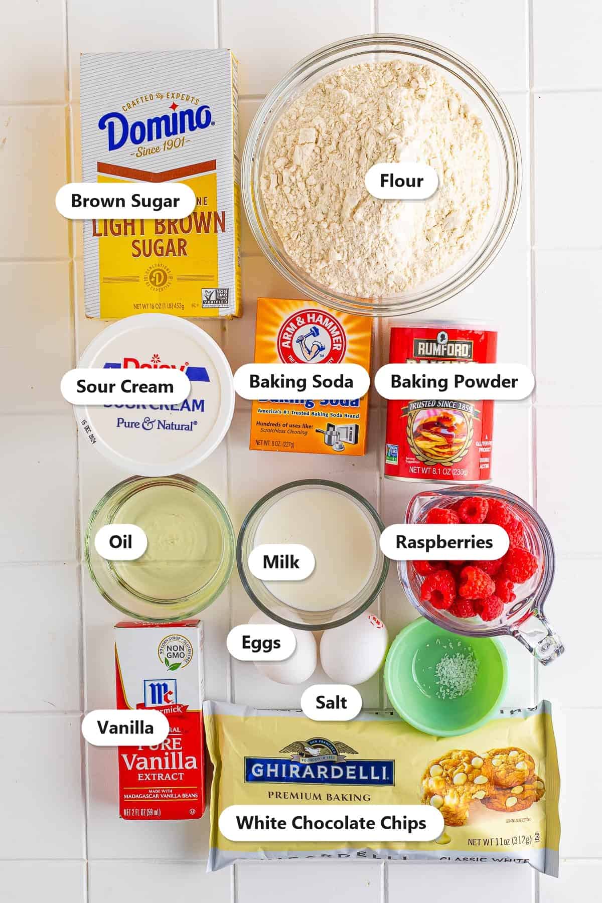 Labeled ingredients for white chocolate raspberry muffins including flour, brown sugar, sour cream, eggs, oil, fresh raspberries, and white chocolate chips.