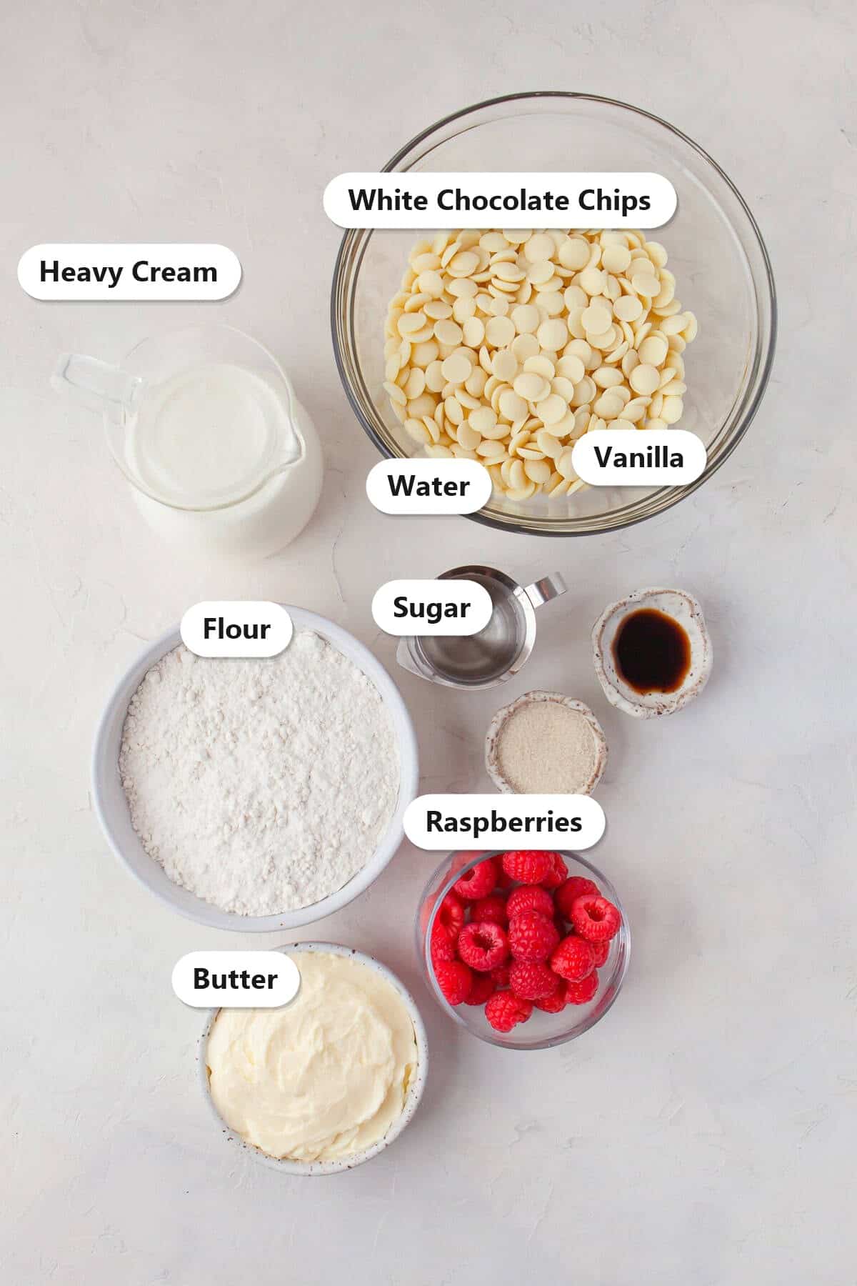 Labeled ingredients to make a white chocolate raspberry tart including white chocolate chips, heavy cream, flour, butter, raspberries, sugar, and vanilla.