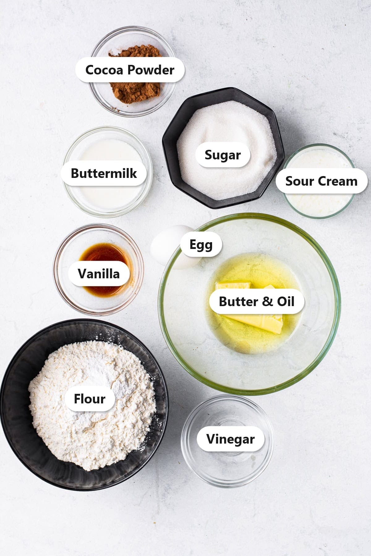 Labeled ingredients for red velvet cupcakes including cocoa powder, sugar, buttermilk, sour cream, egg, butter and oil, vanilla, flour, and vinegar.