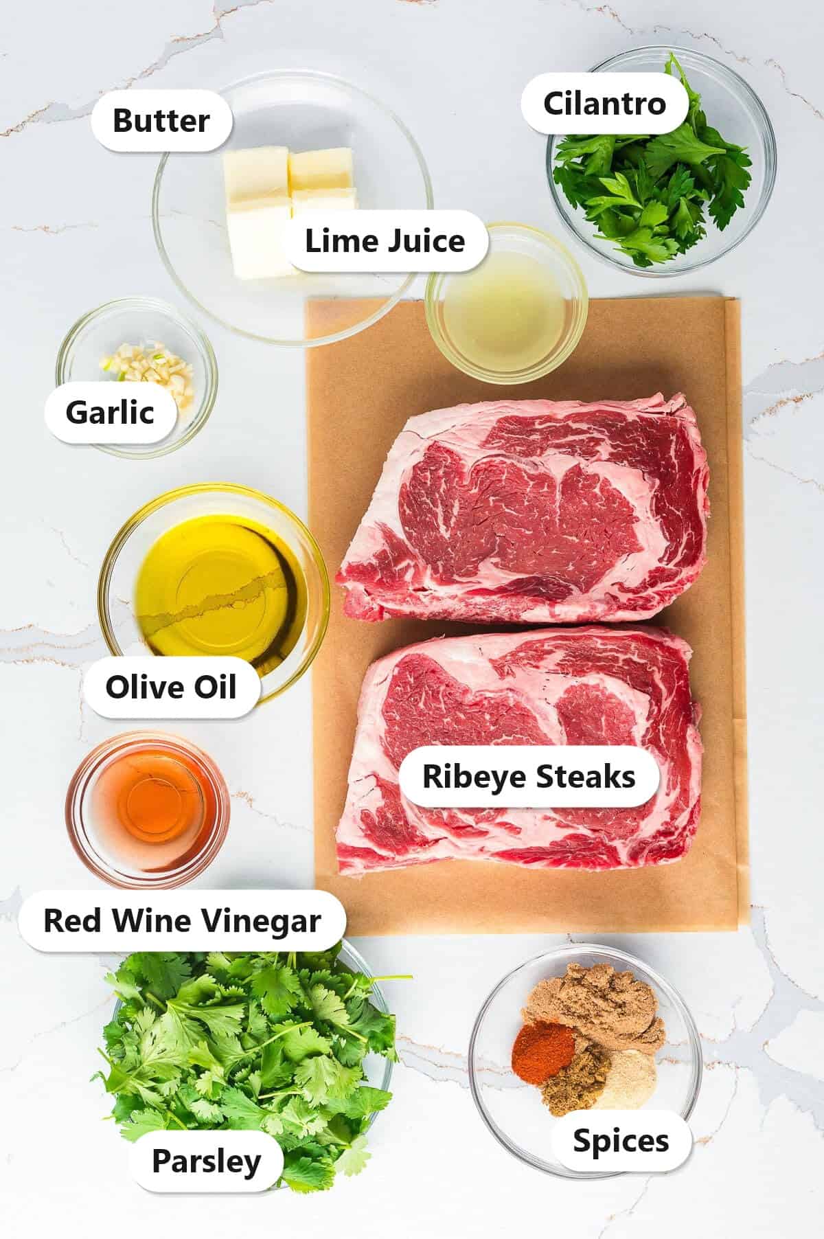 Labeled ingredients for ribeye steak marinade including ribeye steaks, cilantro, parsley, lime, olive oil, garlic, butter, red wine vinegar, and spices.