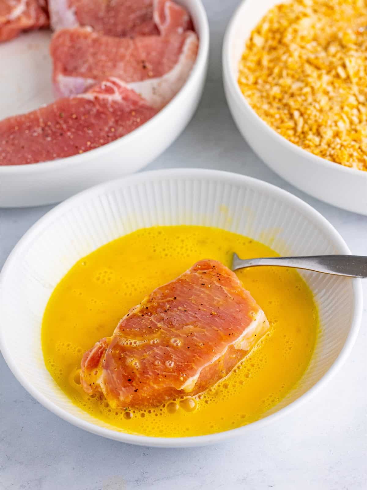 A pork chop being dipped into beaten egg before coating in the Ritz cracker crumbs.