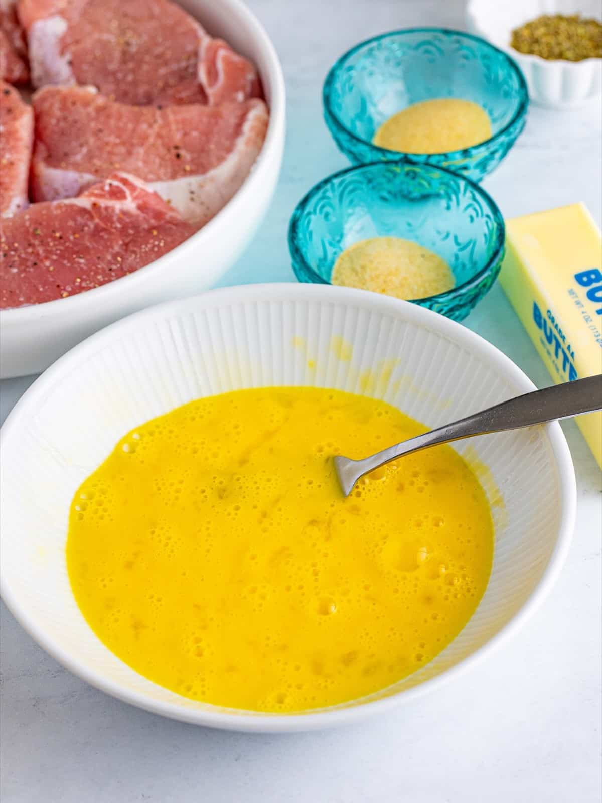 Boneless pork chops seasoned with salt and pepper next to a bowl of beaten eggs for breading.