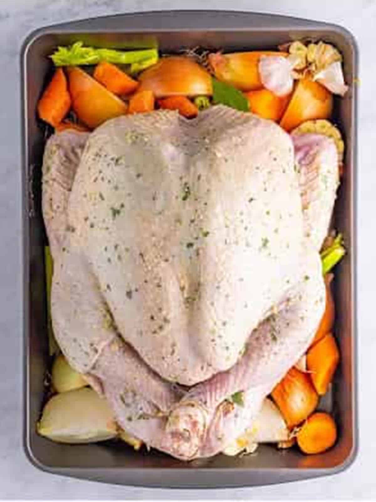 Buttered whole turkey placed on a bed of onions, carrots, and celery in a roasting pan.