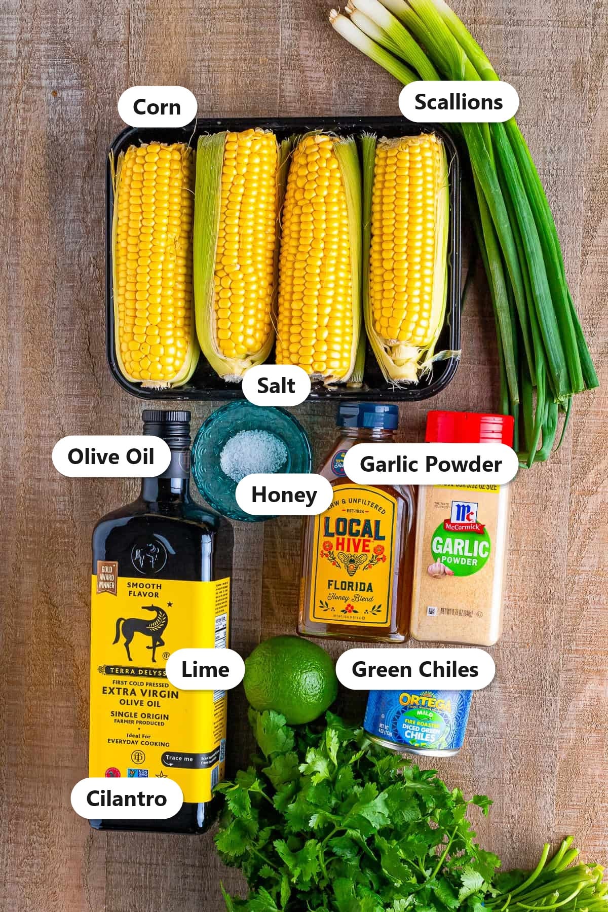 Labeled ingredients for grilled corn salsa including corn, scallions, olive oil, salt, honey, garlic powder, lime, green chiles, and cilantro.