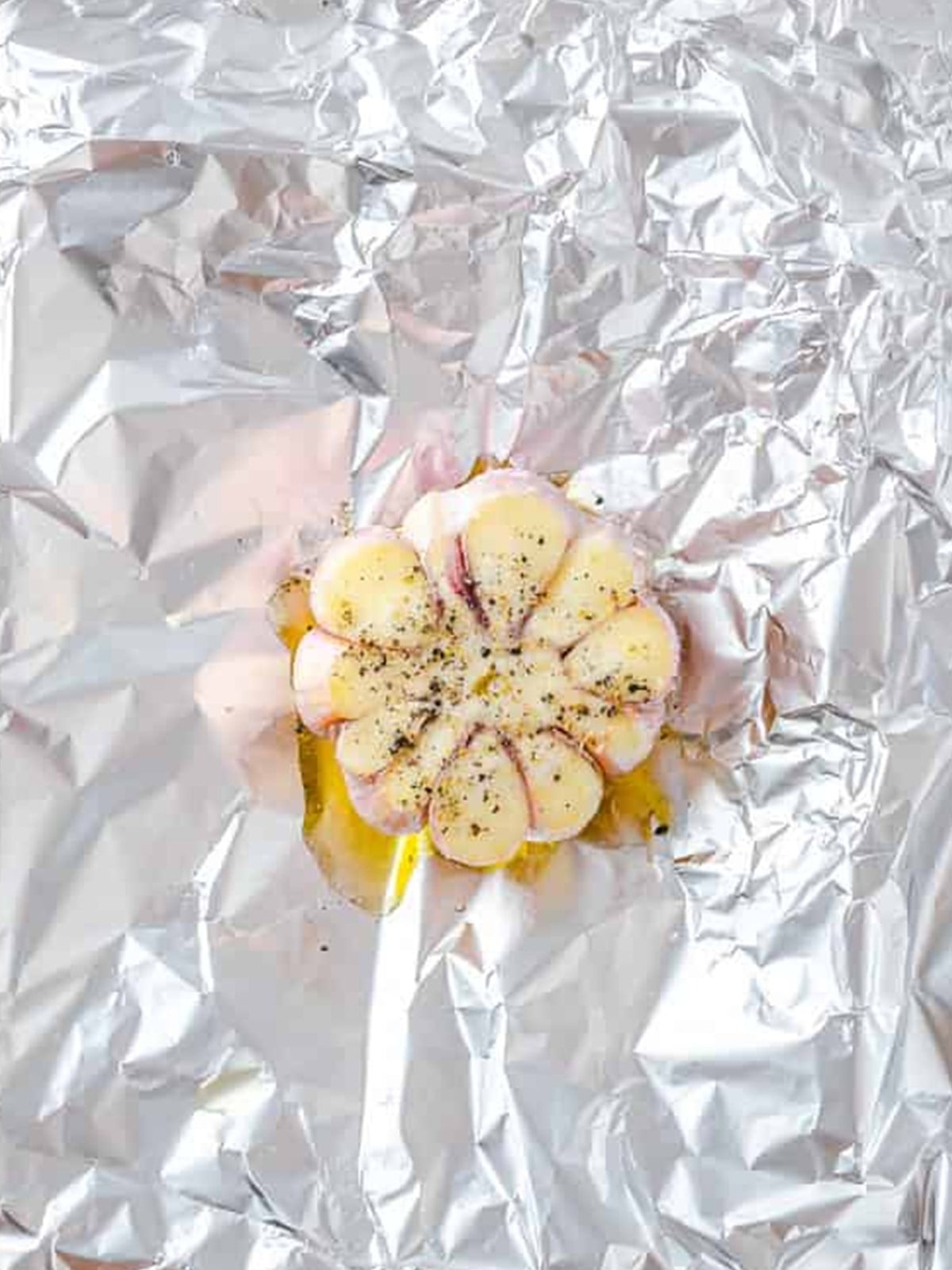 A head of garlic with the top sliced off, drizzled with olive oil and seasoned with salt and pepper on foil.