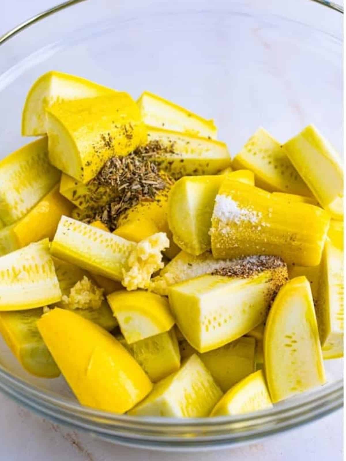 Yellow squash pieces in a glass bowl with olive oil, garlic, herbs de Provence, salt, and pepper.