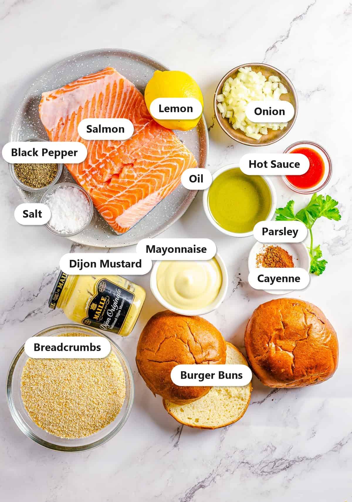 Ingredients to make salmon burgers labeled on a marble surface: salmon, lemon, onion, black pepper, salt, oil, hot sauce, parsley, mayonnaise, dijon mustard, cayenne, breadcrumbs, and burger buns.