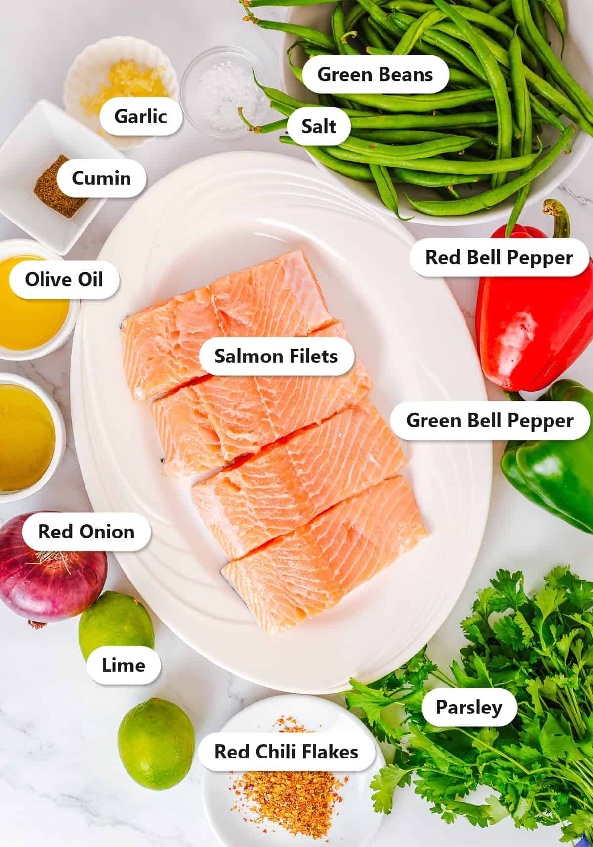 Labeled ingredients for baked salmon sheet pan dinner including salmon filets, green beans, bell peppers, red onion, lime, parsley, olive oil, garlic, cumin, salt, and red chili flakes.