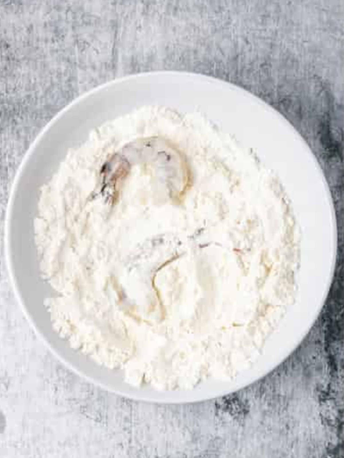Peeled shrimp being dredged in flour on a plate.