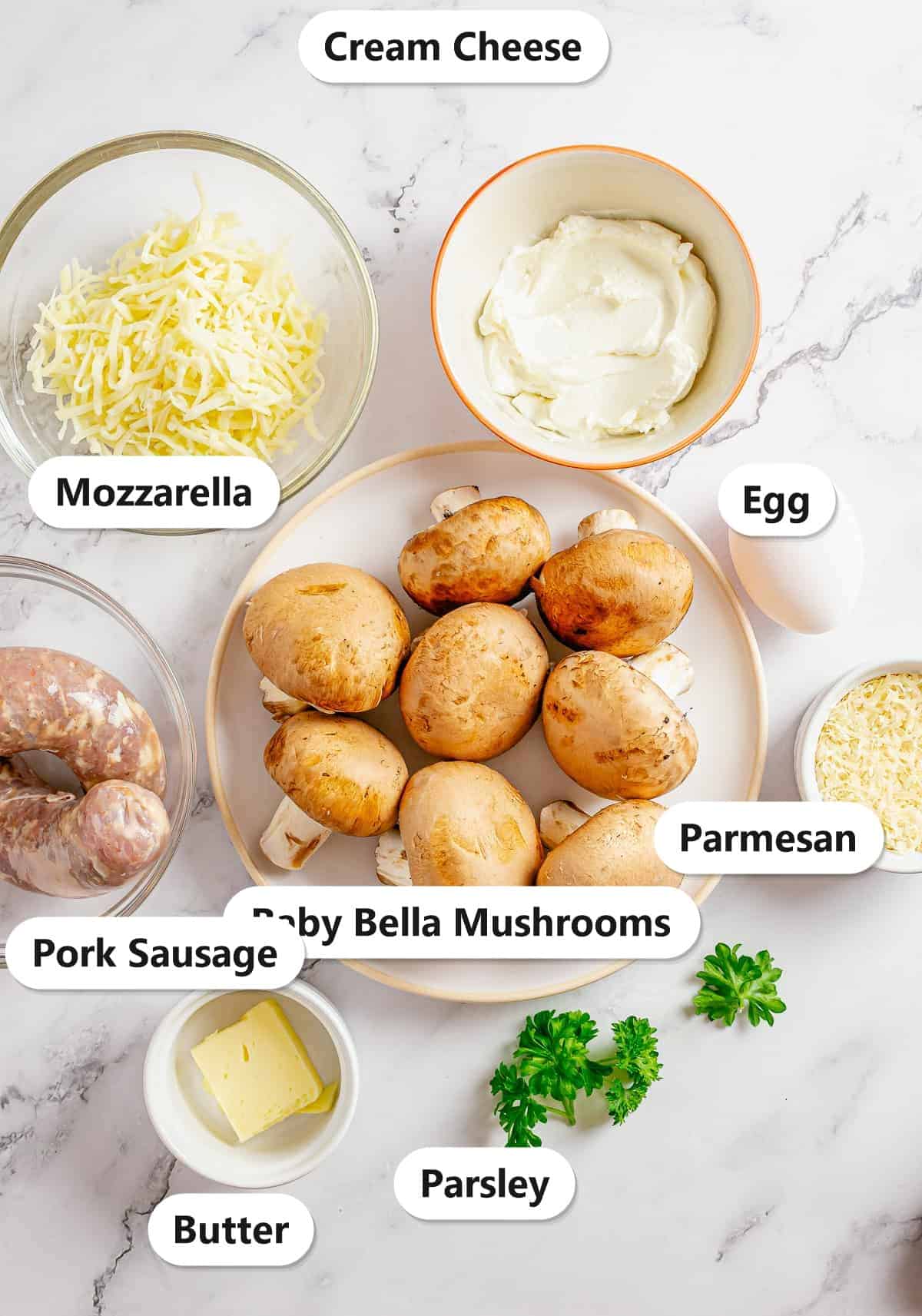 Labeled ingredients for sausage stuffed mushrooms including baby bella mushrooms, pork sausage, cream cheese, mozzarella, parmesan, egg, butter, and parsley.