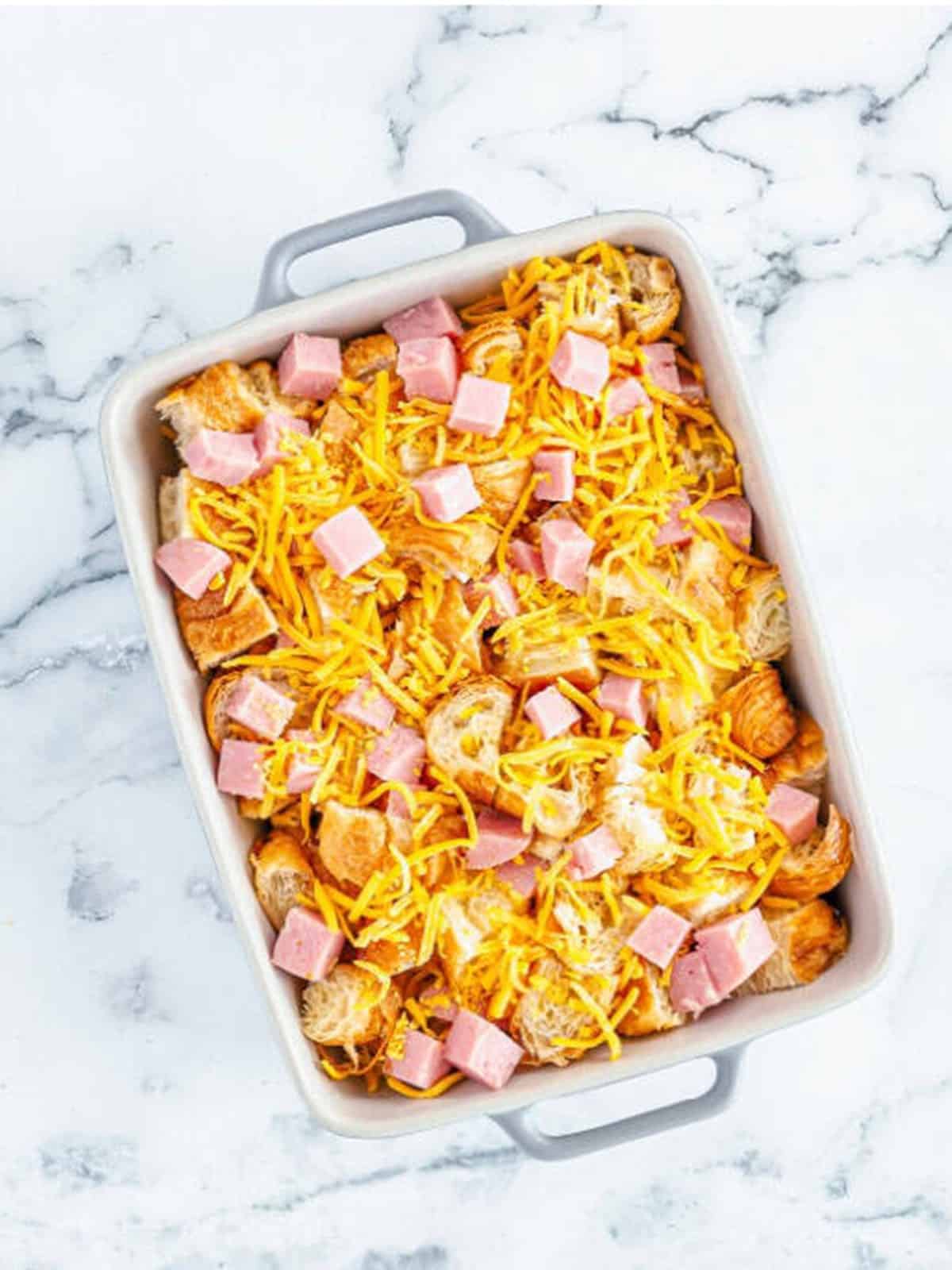 Diced croissants layered with ham and shredded cheddar in a baking dish.