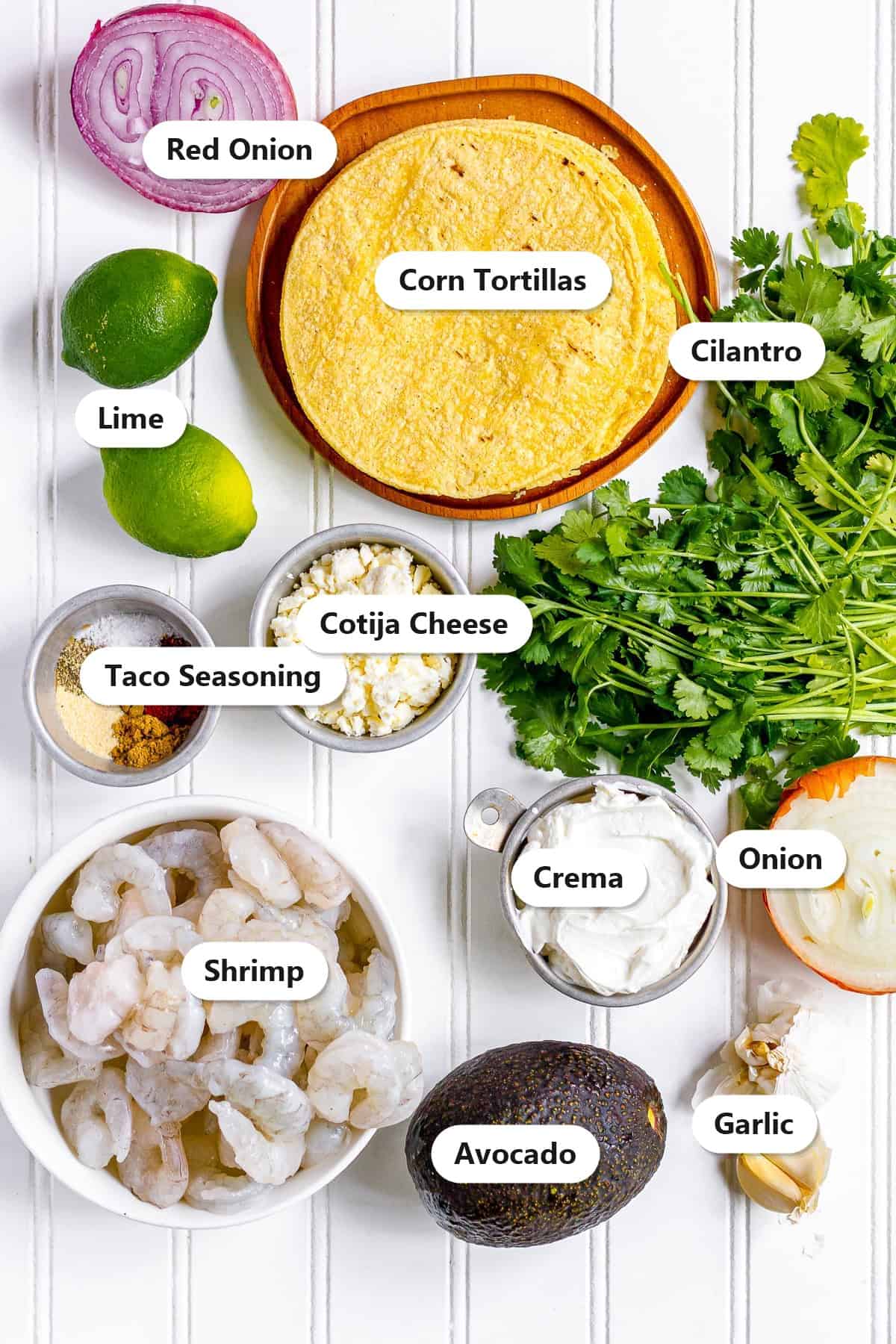 Labeled ingredients for shrimp tacos including shrimp, corn tortillas, cabbage, cotija cheese, avocado, lime, cilantro, red onion, and taco seasoning.