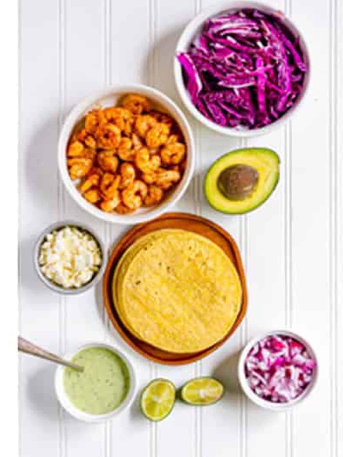 Shrimp taco toppings including cabbage, avocado, cotija, onion, and tortillas laid out.