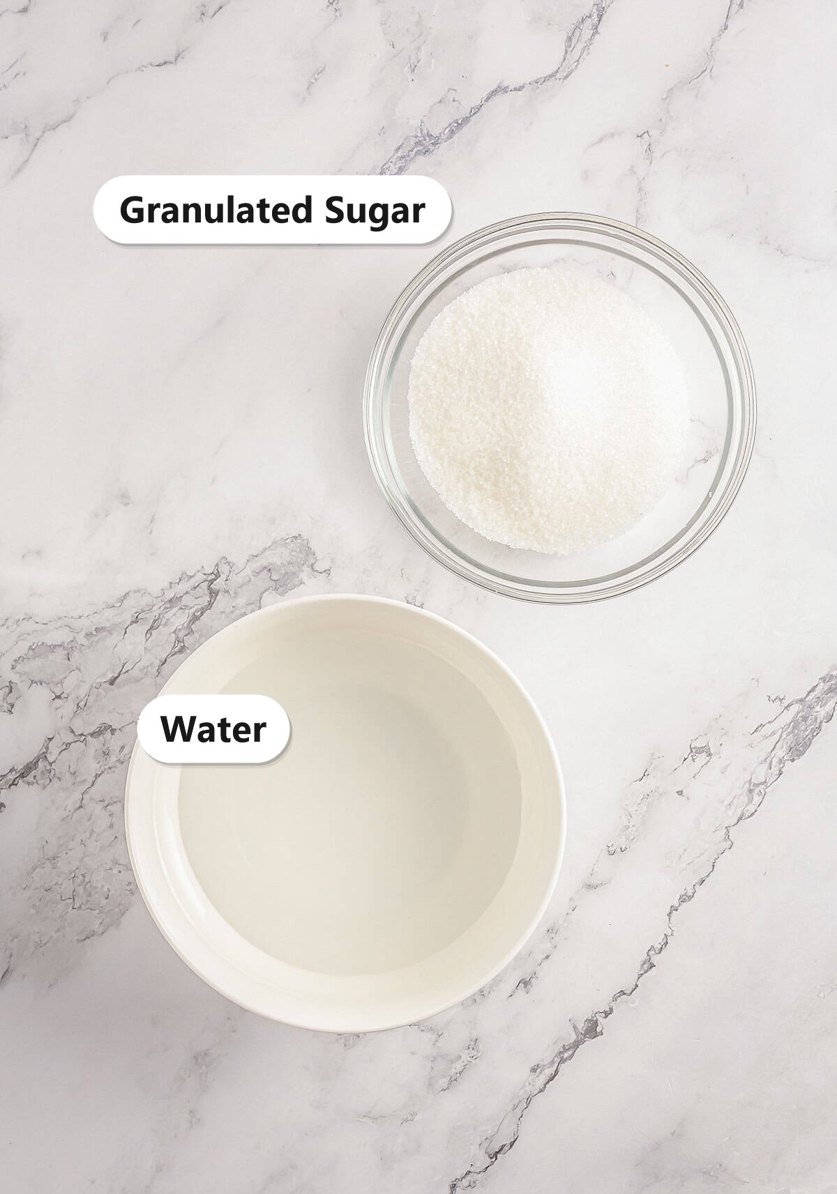 Simple syrup ingredients, granulated sugar and water, labeled in glass bowls on a marble surface.