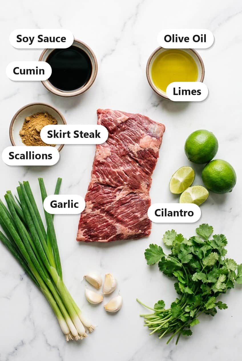 Labeled marinade ingredients for skirt steak tacos including skirt steak, soy sauce, olive oil, cumin, limes, scallions, garlic, and cilantro.