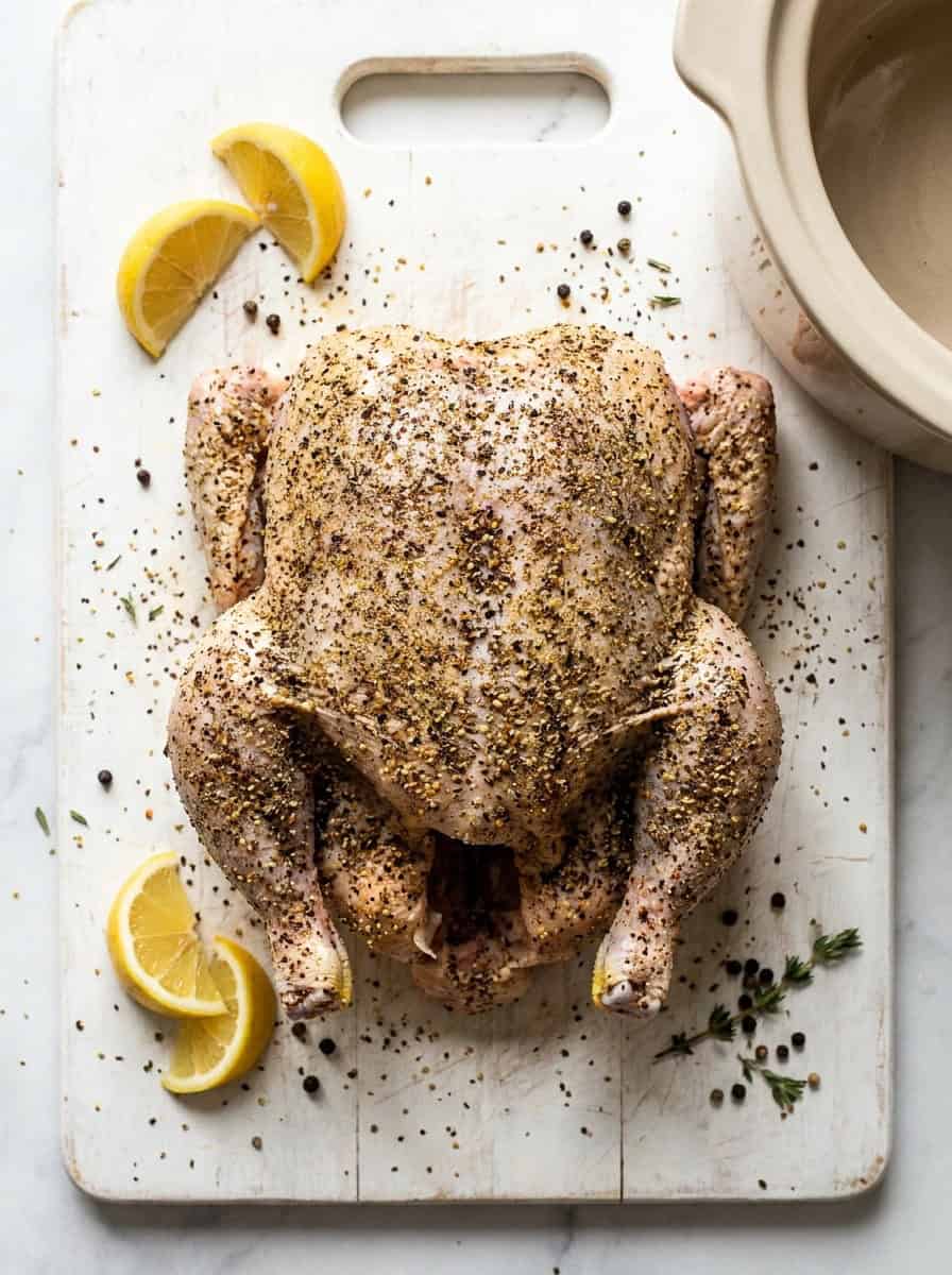 A whole chicken rubbed all over with lemon pepper seasoning on a white cutting board.