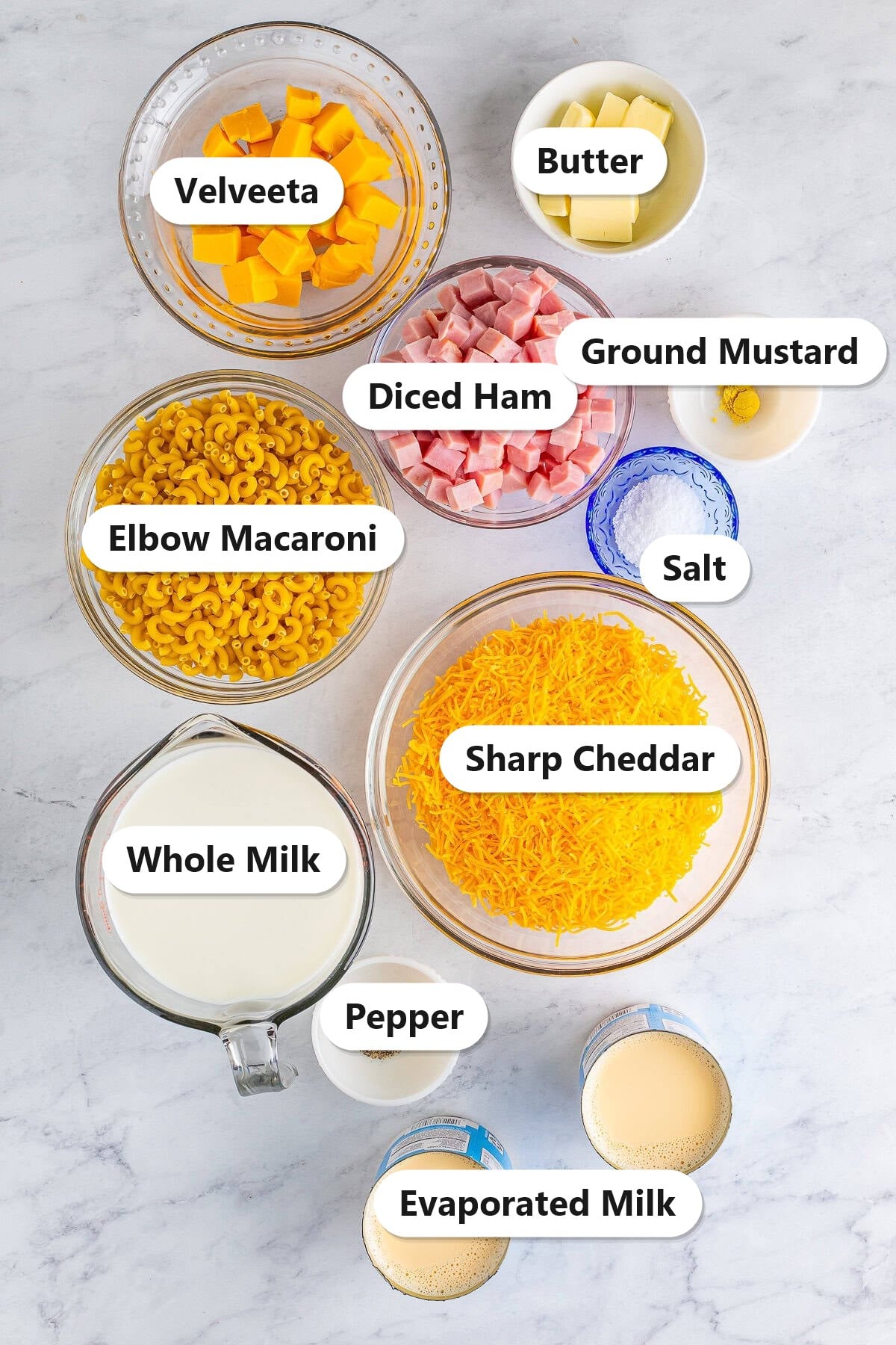 Slow cooker mac and cheese ingredients including elbow macaroni, sharp cheddar, Velveeta, diced ham, whole milk, evaporated milk, and butter labeled on a marble surface.