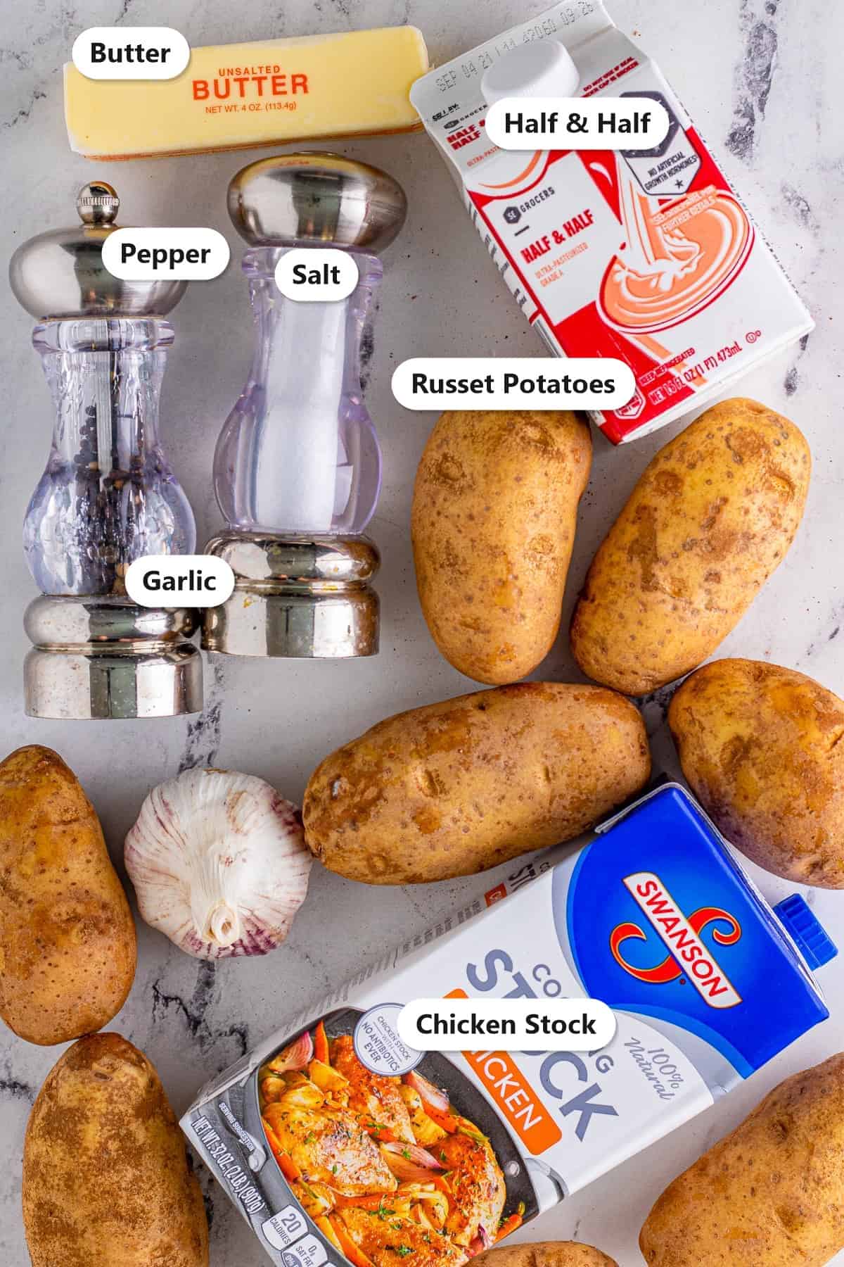 Slow cooker mashed potatoes ingredients labeled on marble including russet potatoes, garlic, butter, half and half, chicken stock, salt, and pepper.