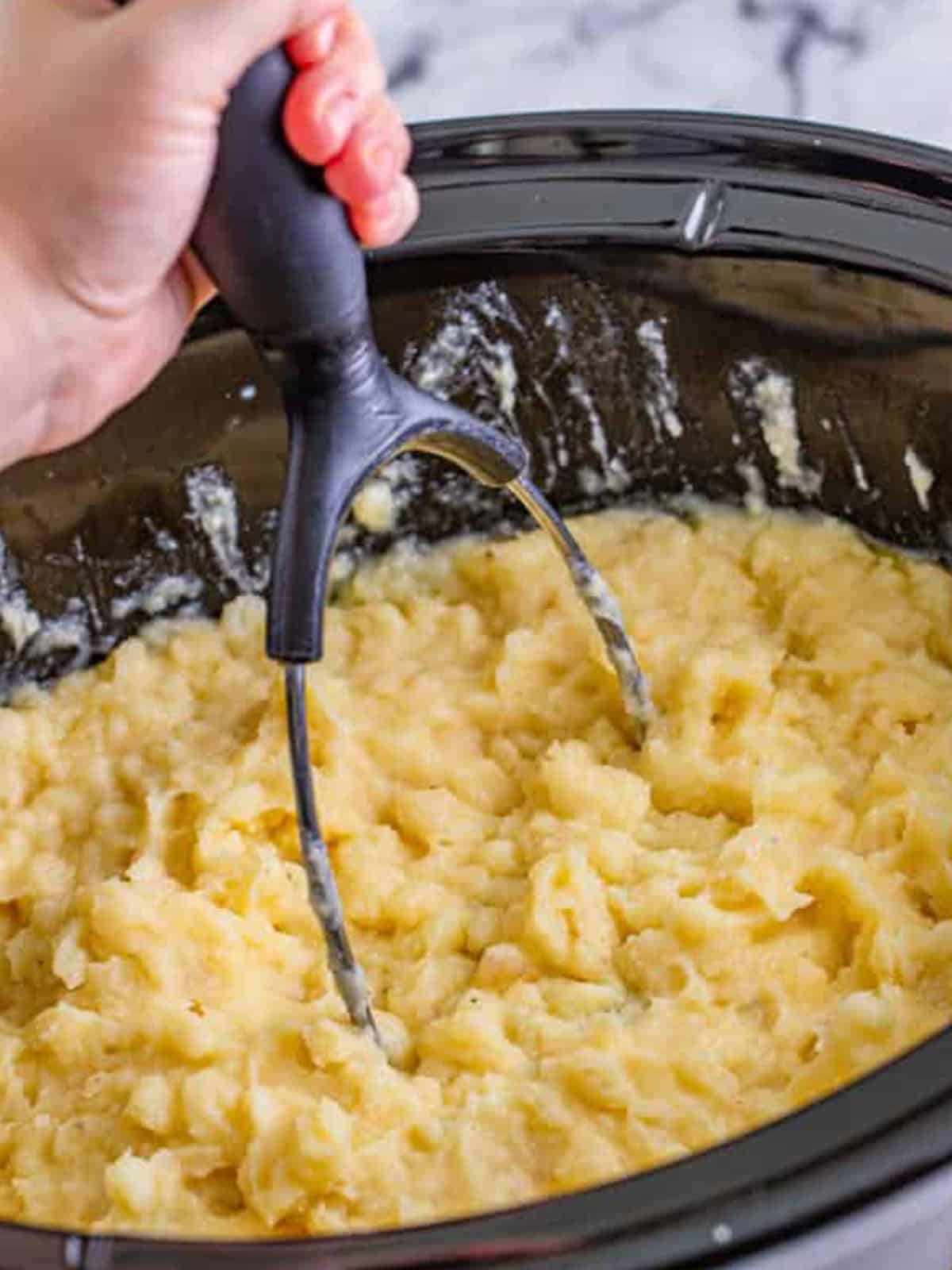 Mashing the cooked potatoes into creamy mashed potatoes with a potato masher in the slow cooker.