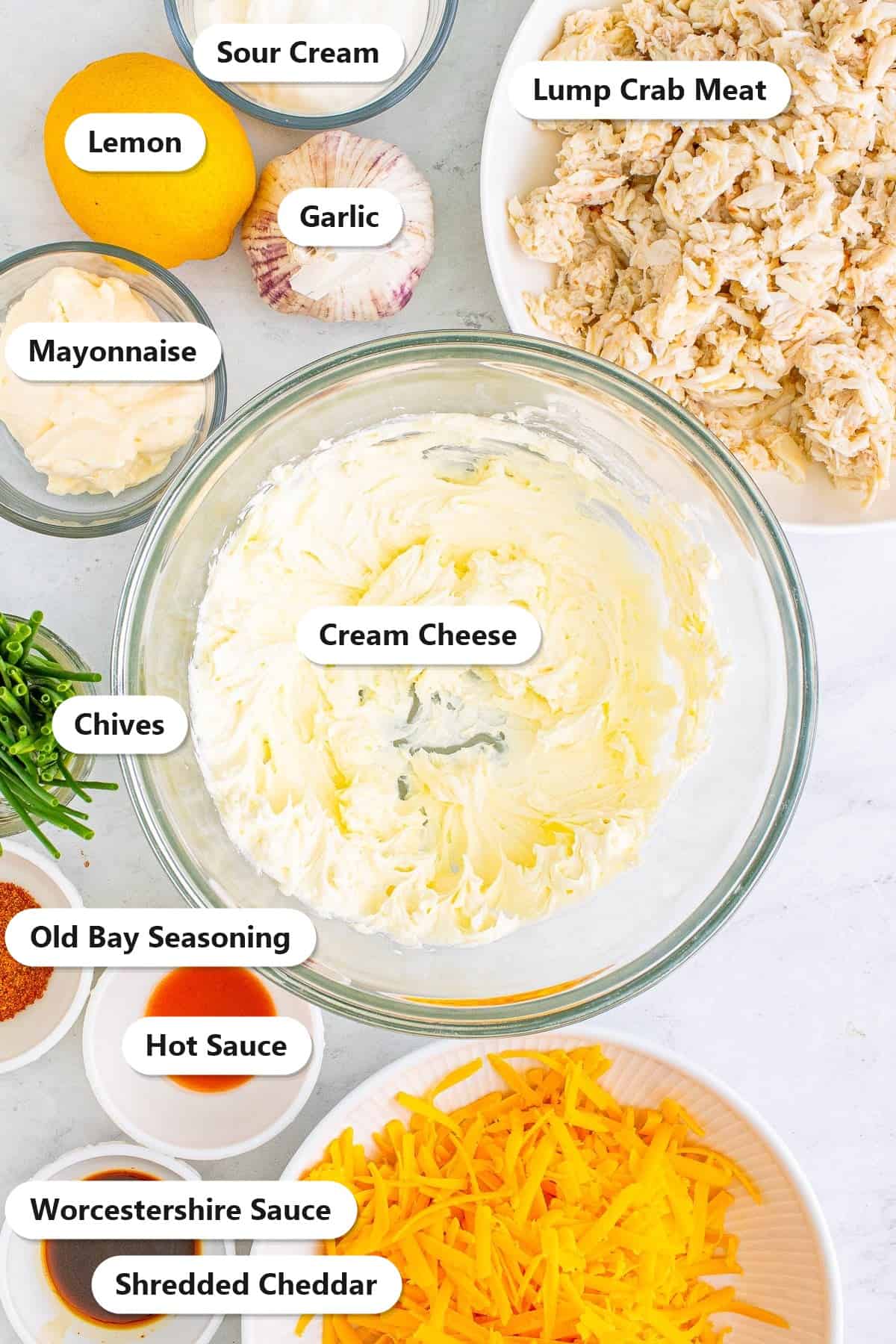 Labeled ingredients for Smoked Crab Dip including cream cheese, sour cream, mayonnaise, lump crab meat, shredded cheddar, Old Bay seasoning, hot sauce, Worcestershire sauce, chives, lemon, and garlic.