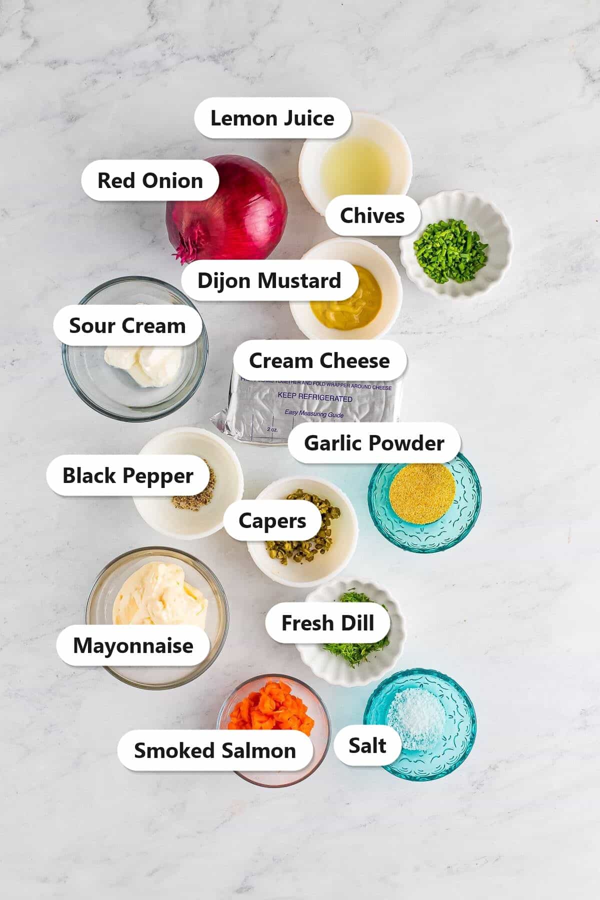 Smoked salmon dip ingredients including cream cheese, smoked salmon, mayonnaise, sour cream, capers, dill, chives, and red onion labeled on a marble surface.