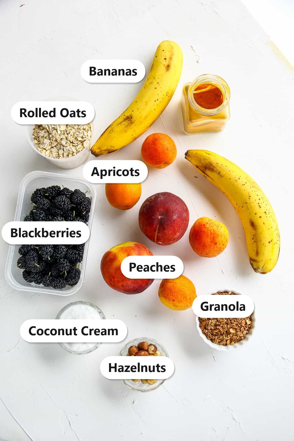 Labeled ingredients to make a smoothie bowl including bananas, apricots, peaches, rolled oats, coconut cream, blackberries, granola, and hazelnuts.