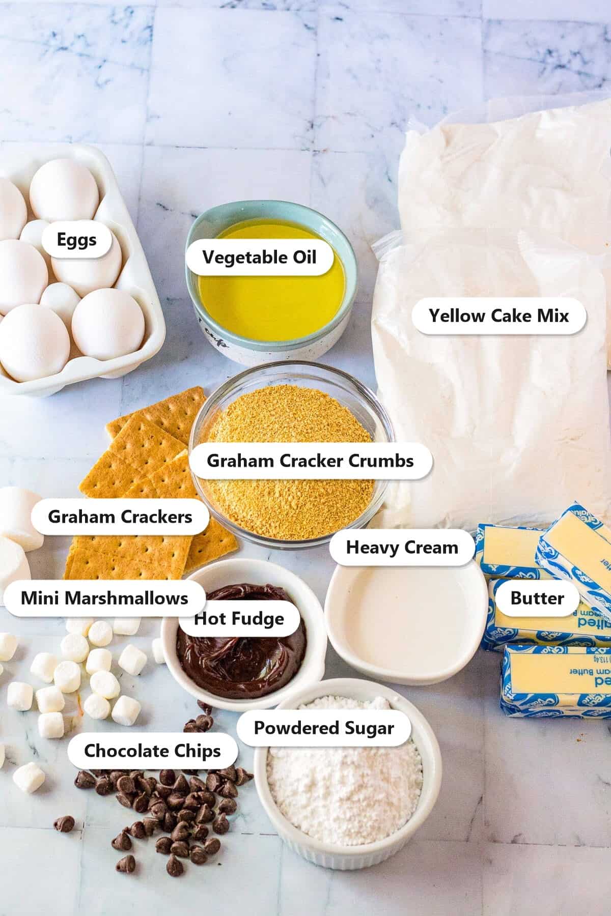 Ingredients to make a s'mores cake including yellow cake mix, graham cracker crumbs, eggs, oil, hot fudge, butter, marshmallows, chocolate chips, and heavy cream.