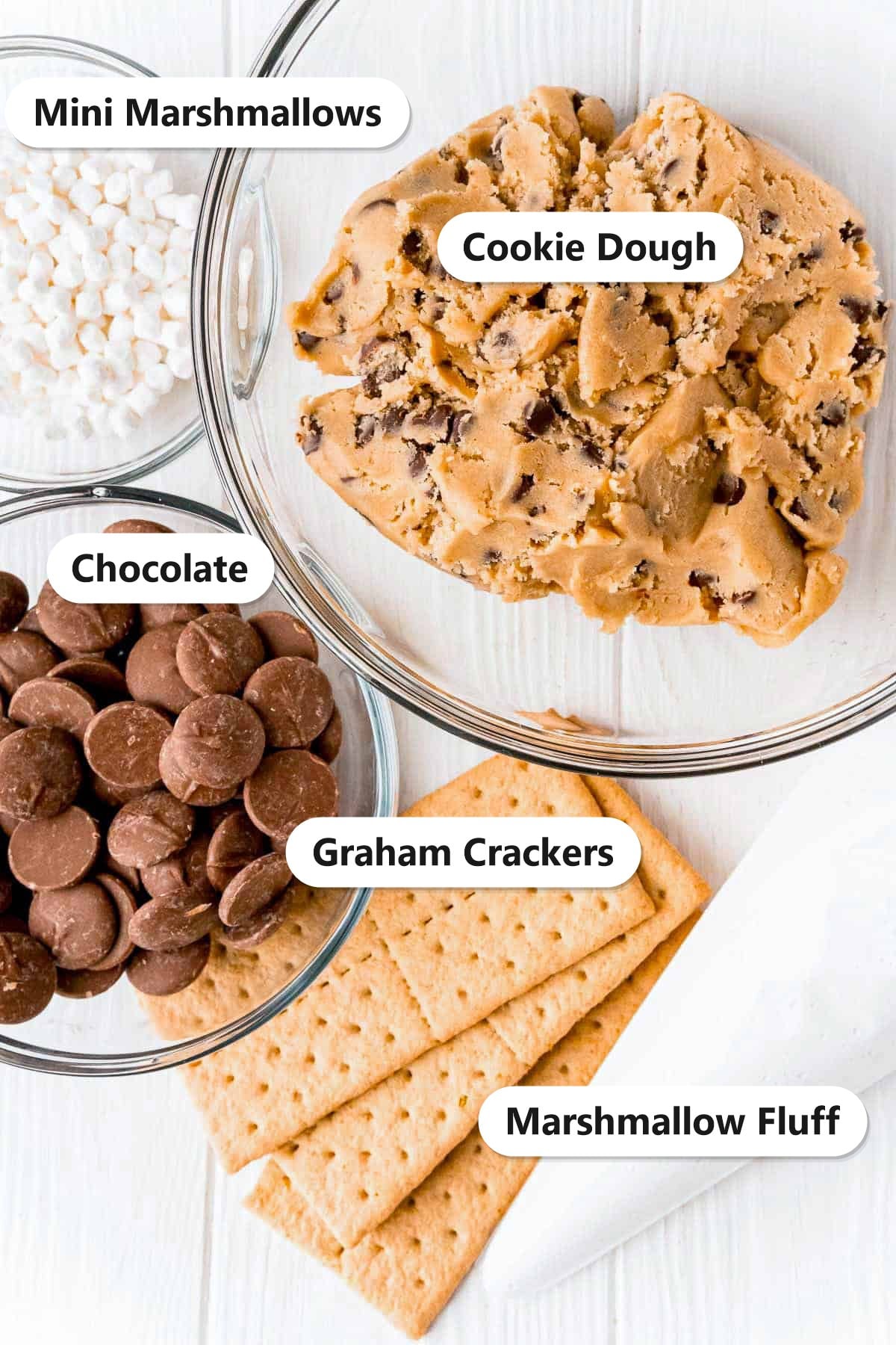 Ingredients for s'mores cookies including cookie dough, mini marshmallows, chocolate, graham crackers, and marshmallow fluff.