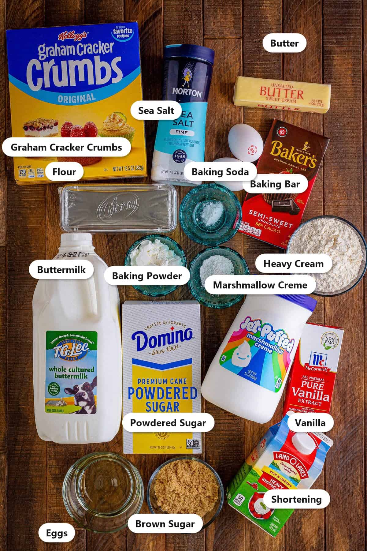 Labeled ingredients for smores whoopie pies including graham cracker crumbs, flour, butter, brown sugar, eggs, buttermilk, baking bar, heavy cream, shortening, powdered sugar, marshmallow creme, and vanilla.