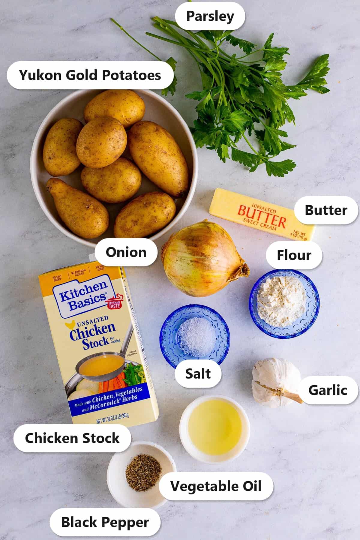 Labeled ingredients for smothered potatoes including yukon gold potatoes, onion, garlic, flour, butter, chicken stock, vegetable oil, parsley, salt, and black pepper.