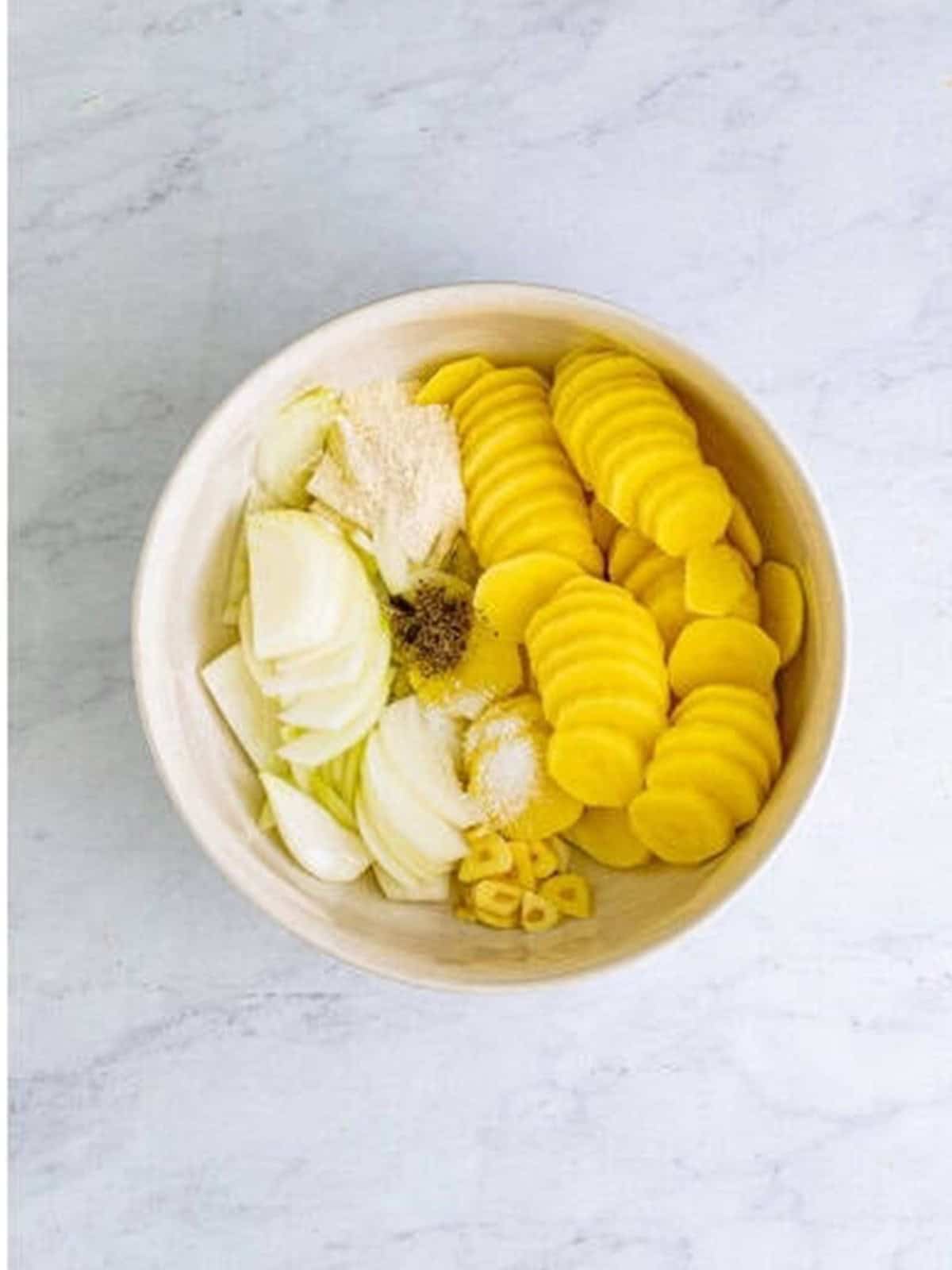 Sliced yukon gold potatoes in a bowl with onion, garlic, flour, salt, and pepper for smothered potatoes.