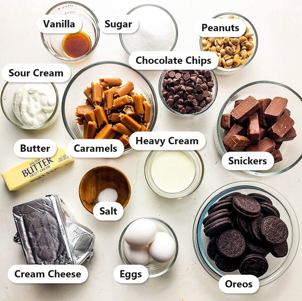 Labeled ingredients for Snickers cheesecake including Oreos, cream cheese, Snickers bars, caramels, peanuts, chocolate chips, eggs, sour cream, and butter.