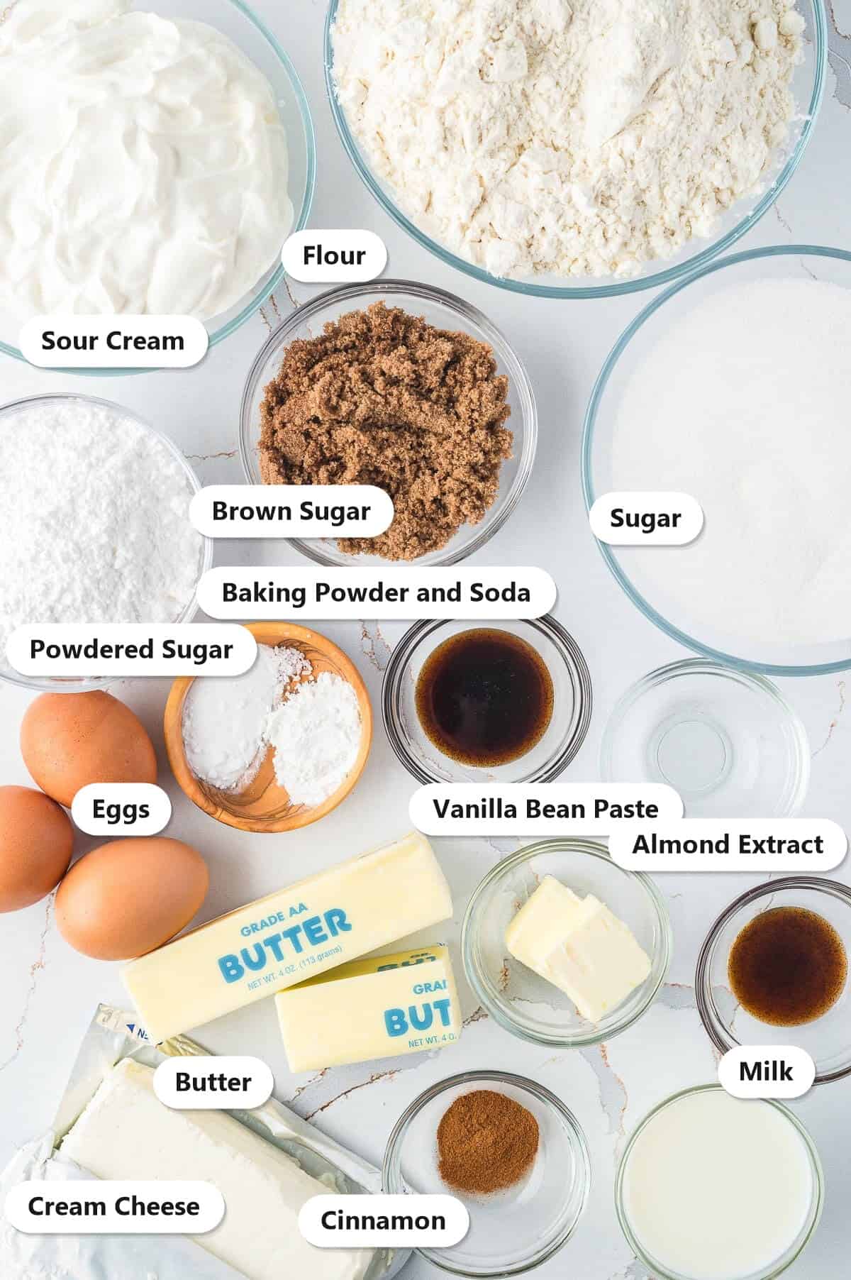 Labeled ingredients for sour cream coffee cake including flour, sour cream, butter, sugar, brown sugar, eggs, cream cheese, powdered sugar, cinnamon, vanilla bean paste, almond extract, and milk.