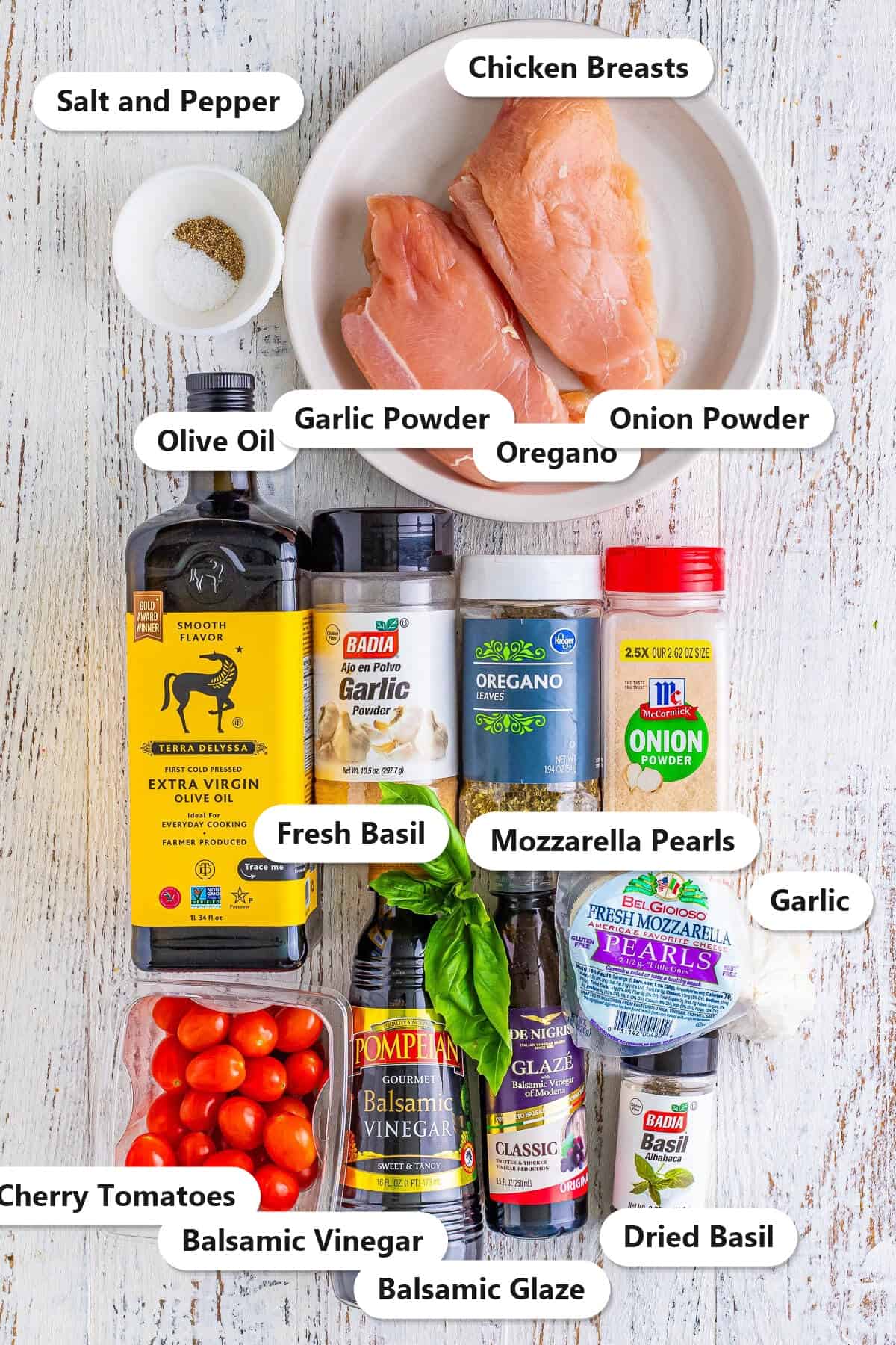 Labeled ingredients to make sous vide chicken breast caprese including chicken breasts, olive oil, garlic powder, oregano, onion powder, dried and fresh basil, cherry tomatoes, balsamic vinegar, balsamic glaze, mozzarella pearls, garlic, salt, and pepper.