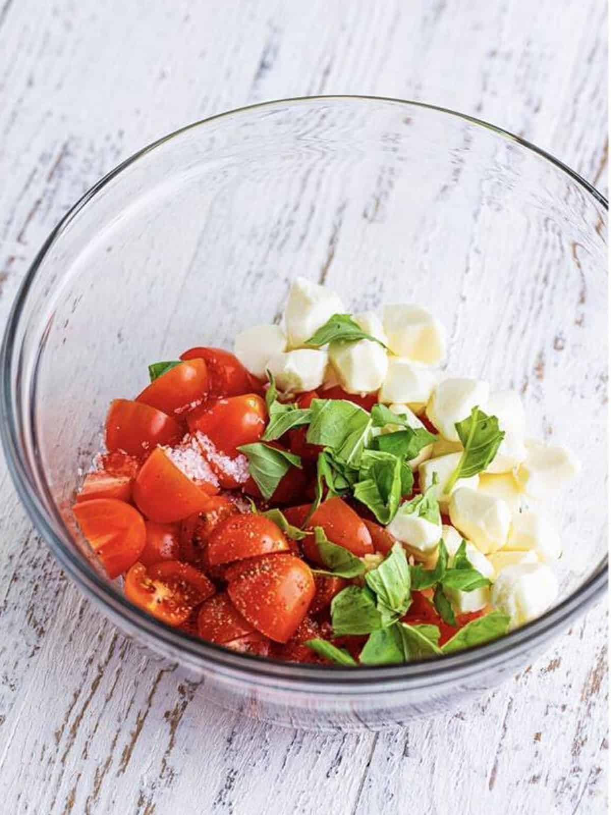 Cherry tomatoes, mozzarella pearls, and fresh basil for the caprese topping in a glass bowl.