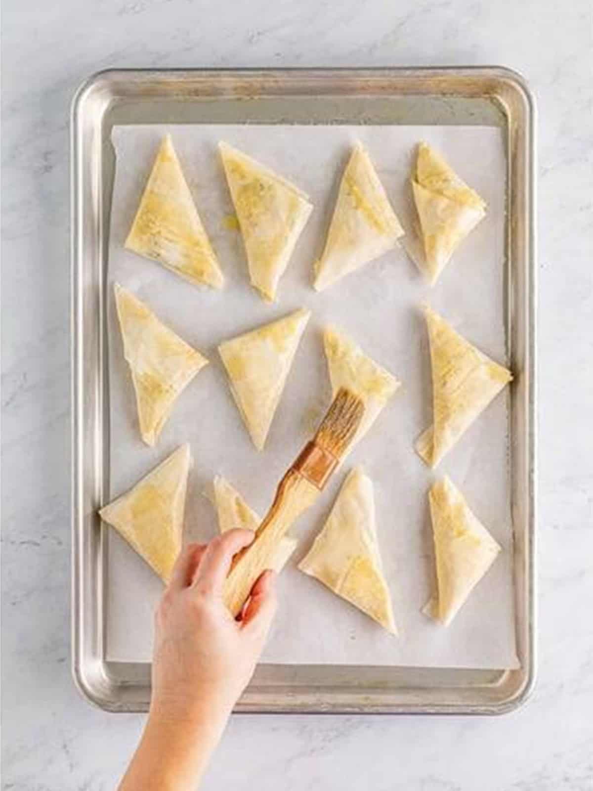 Spanakopita triangles arranged on a tray and brushed with butter.