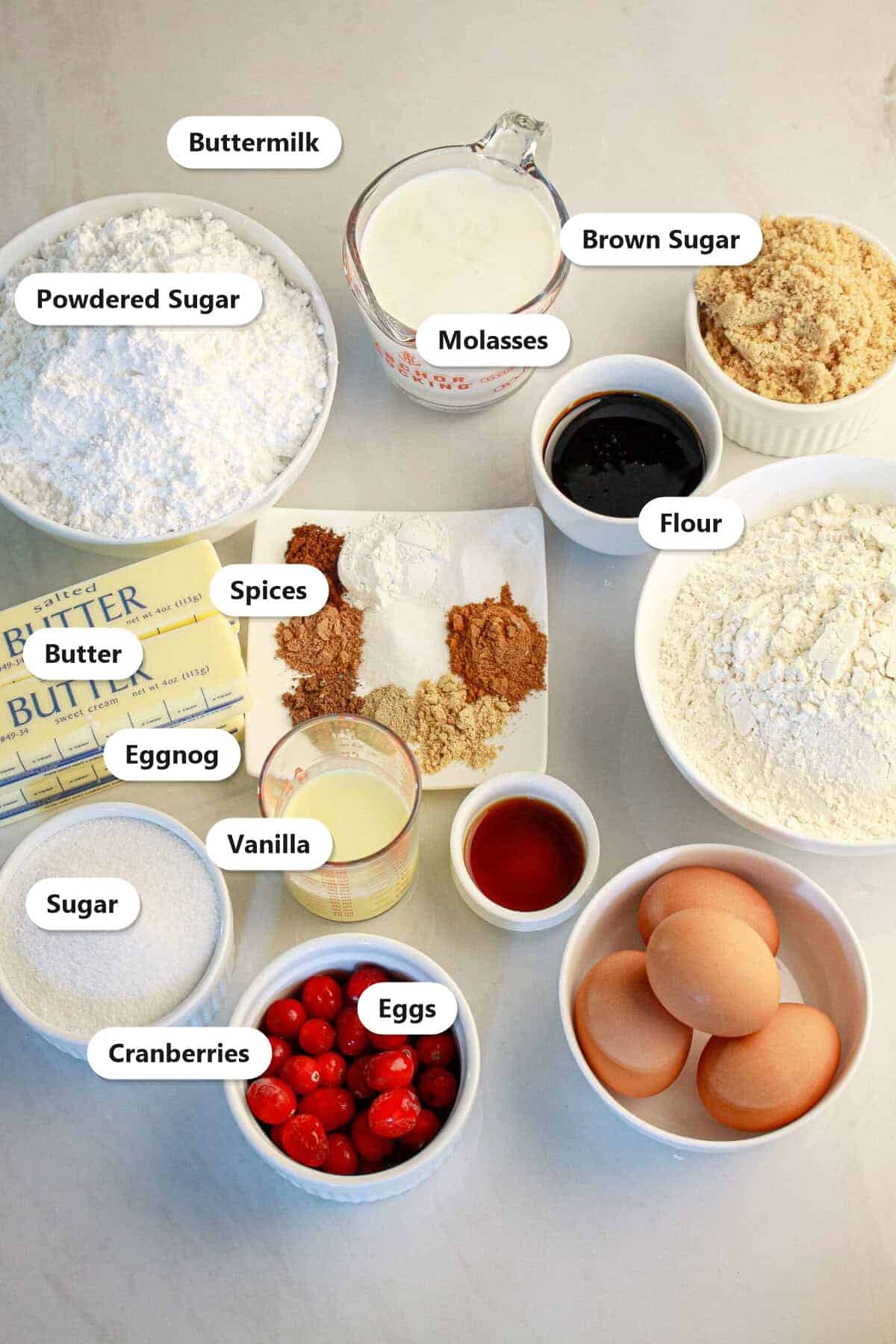 Ingredients to make spice cake including flour, butter, brown sugar, molasses, buttermilk, eggs, warm spices, eggnog, cranberries, and powdered sugar.