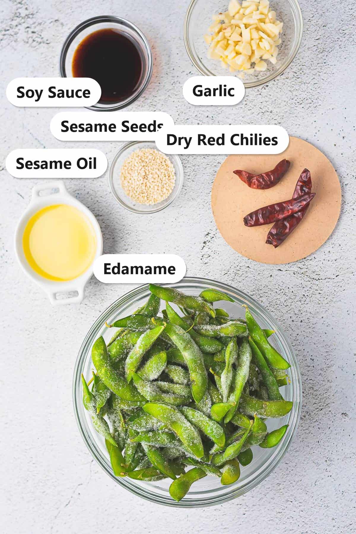 Edamame recipe ingredients including frozen edamame, sesame oil, garlic, dry red chilies, soy sauce, and sesame seeds labeled on a counter.