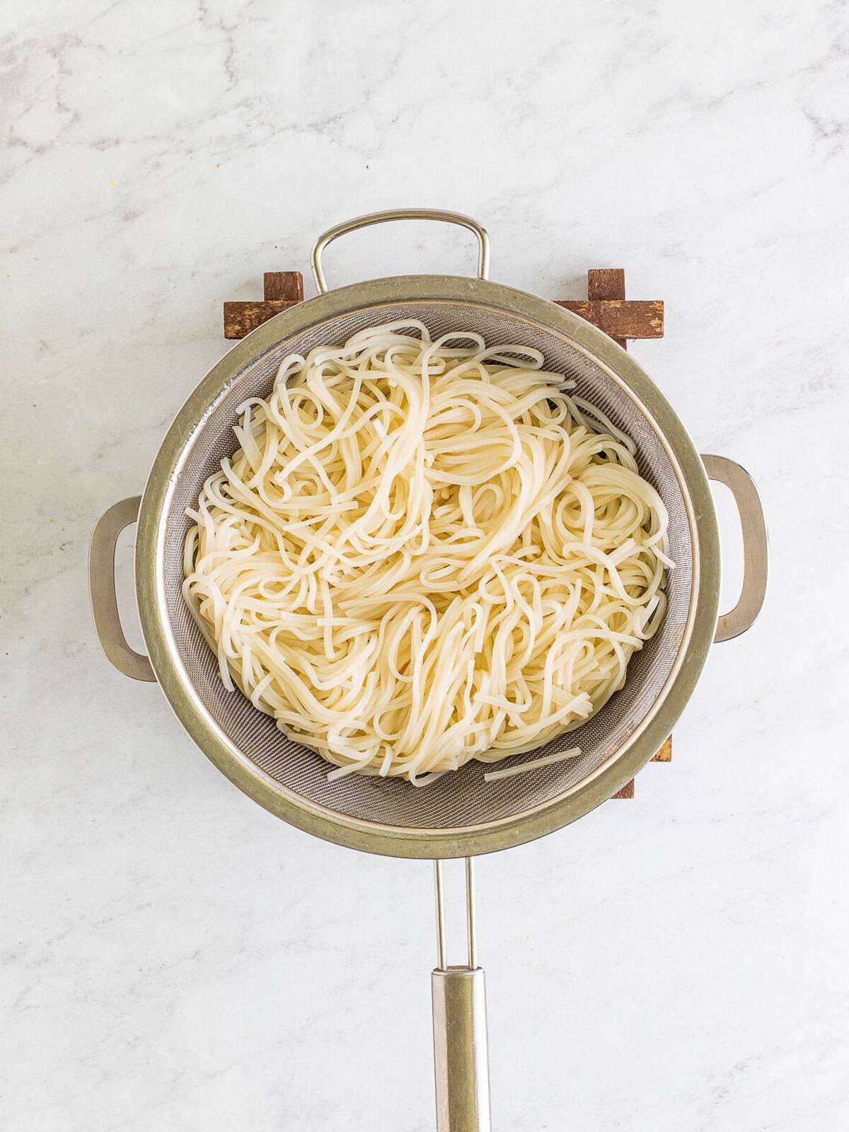 Cooked flat rice noodles draining in a strainer after a cold water rinse.