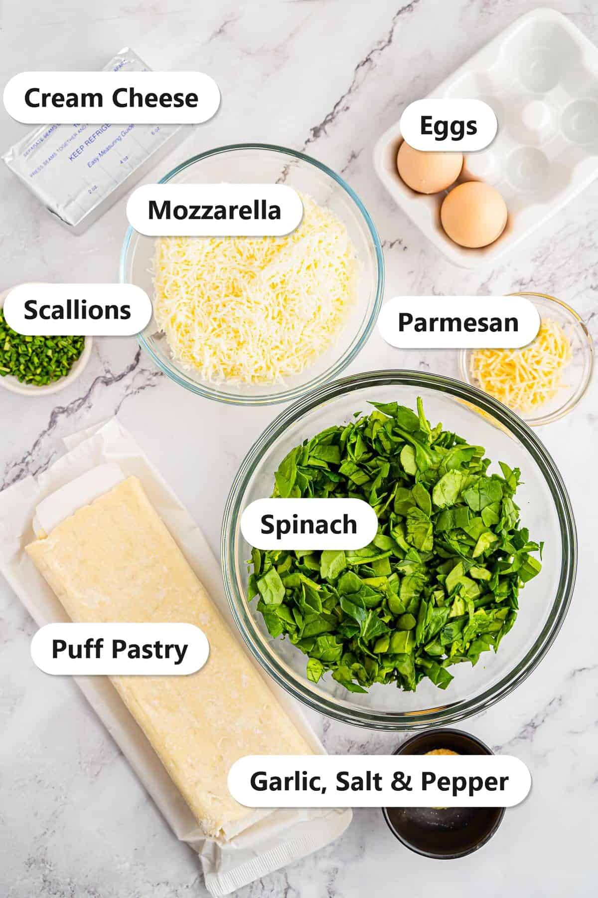 Labeled ingredients to make spinach puffs including cream cheese, mozzarella, Parmesan, fresh spinach, puff pastry, eggs, scallions, garlic powder, salt, and pepper.